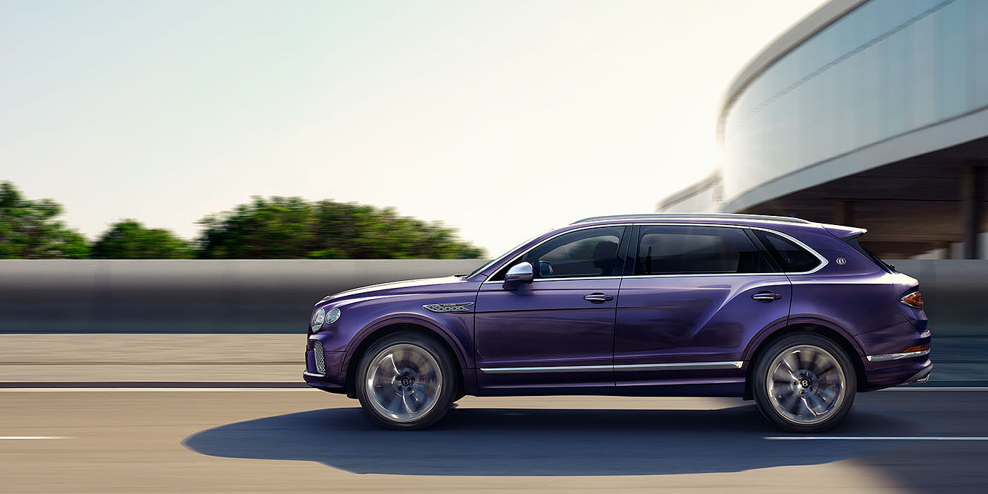 Bentley San Juan Bentley Bentayga Extended Wheelbase Mulliner luxury SUV profile in Tanzanite Purple paint, driving on an country road past trees.